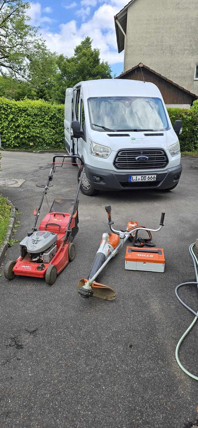 White Ford Transit van parked next to outdoor power tools including a red lawn mower, string trimmer, and hedge trimmer on a driveway