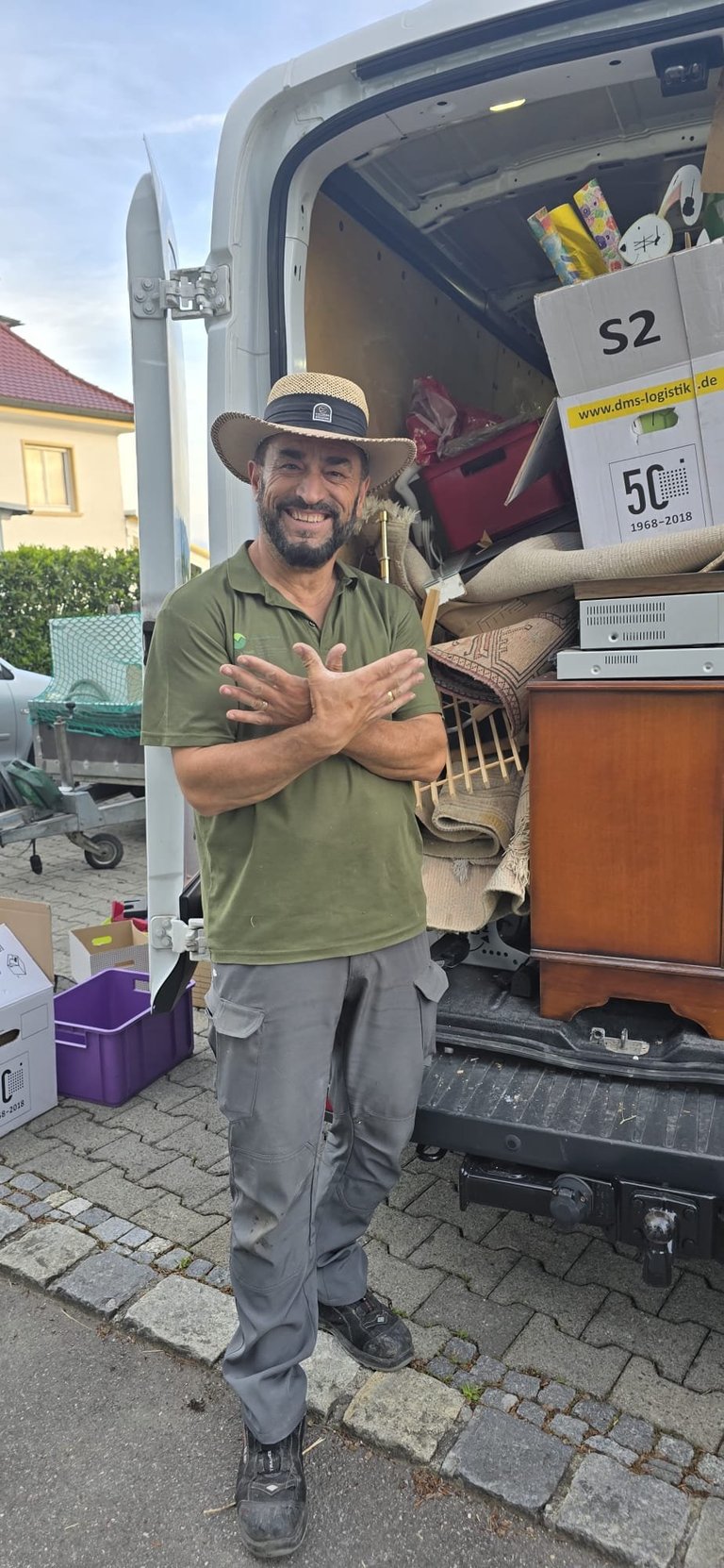 Man in straw hat and olive shirt standing in front of open moving van, smiling with arms crossed, loaded with household items