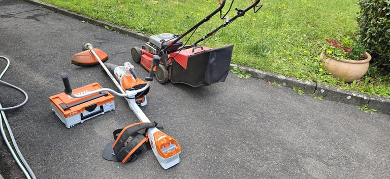 Outdoor power tools including lawn mower, chain saw, and other equipment laid out on asphalt near grass and garden pots.